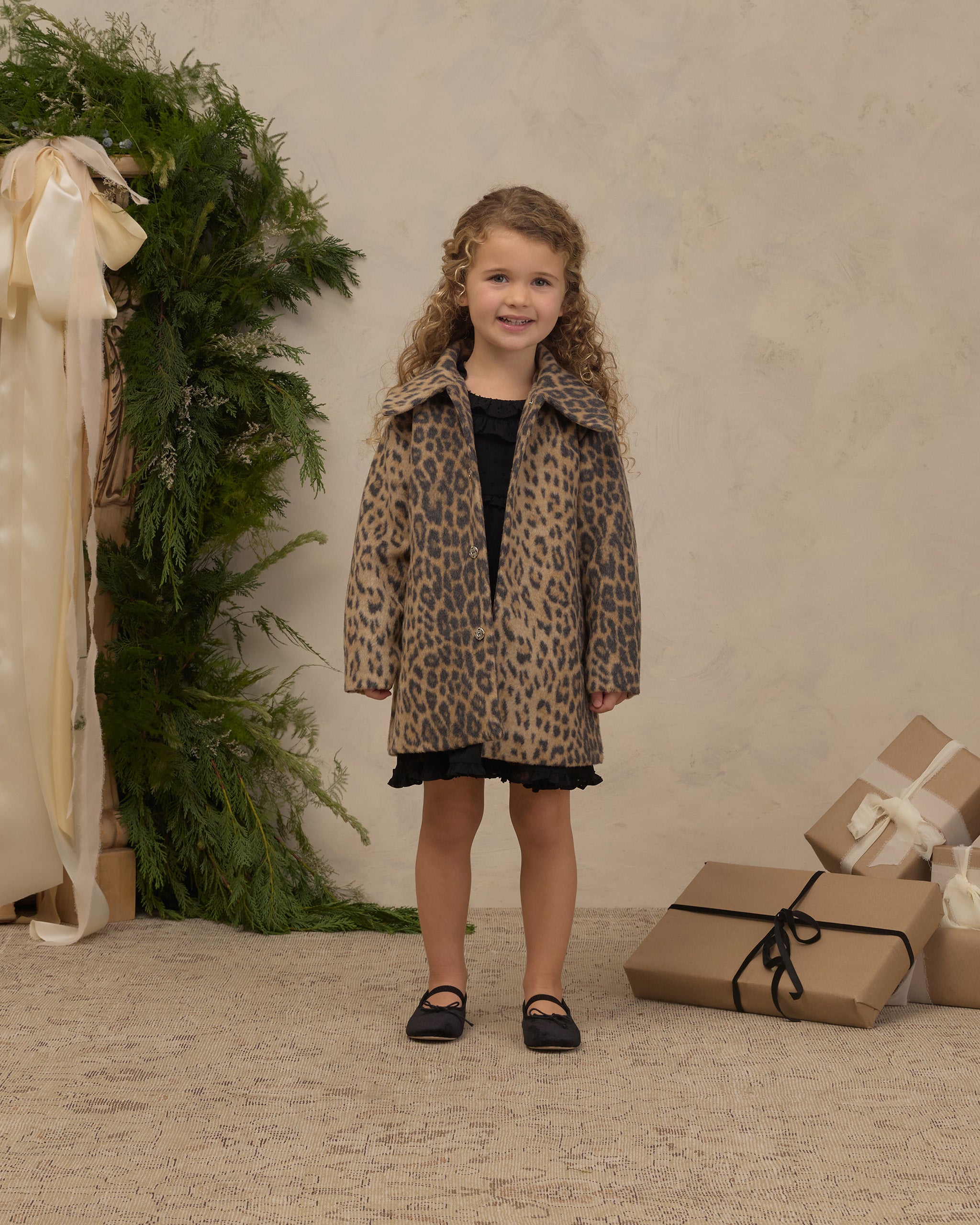 Young girl wearing a leopard print faux fur coat with a full collar and pockets while standing indoors beside holiday greenery and wrapped gifts by Noralee