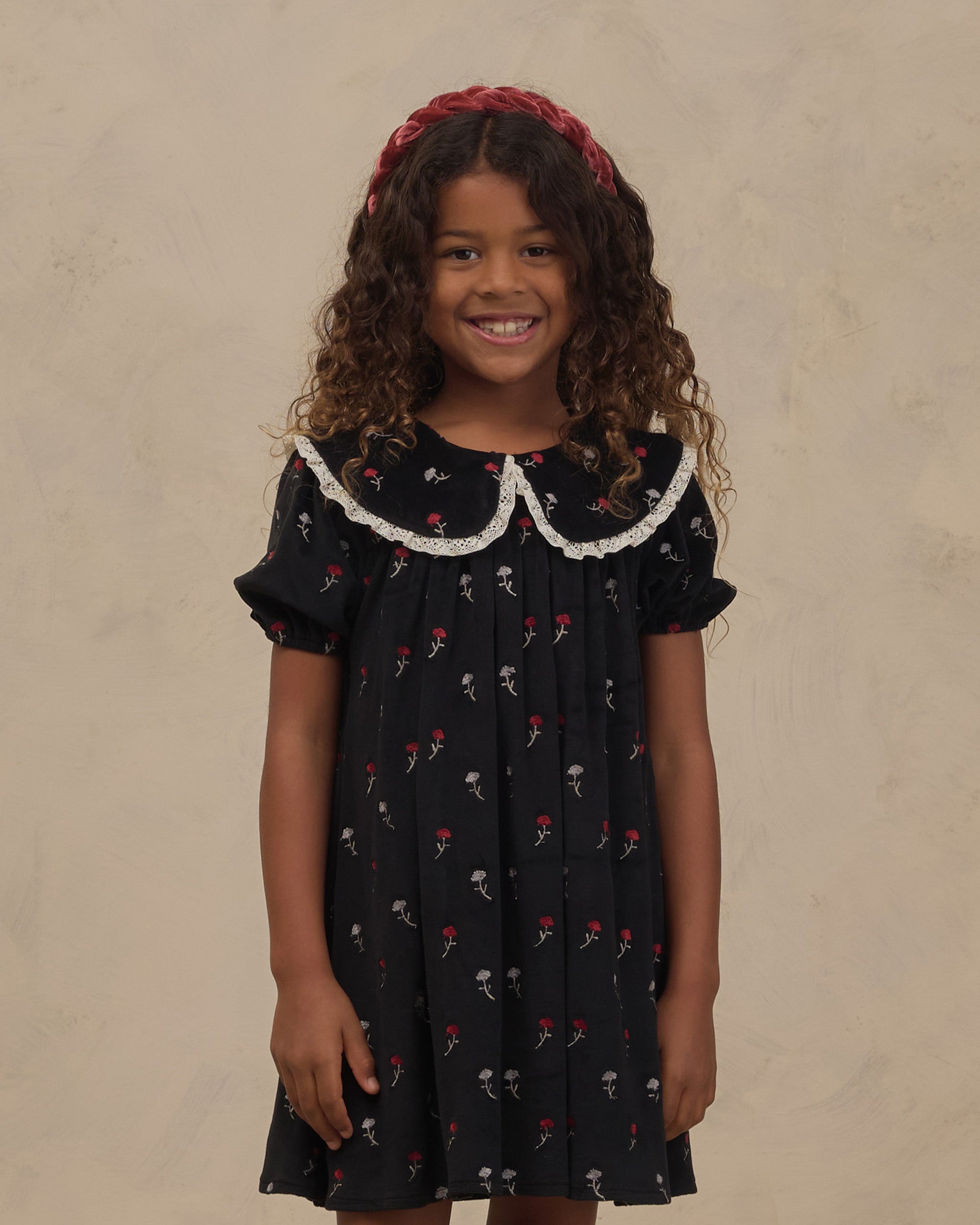 Young girl wearing a black floral dress with a scalloped lace collar and puff sleeves from Noralee with a red headband against a neutral studio backdrop