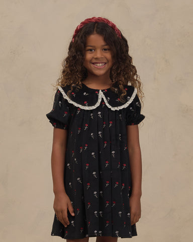 Young girl wearing a black floral dress with a scalloped lace collar and puff sleeves from Noralee with a red headband against a neutral studio backdrop