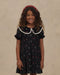 Young girl wearing a black floral dress with a scalloped lace collar and puff sleeves from Noralee with a red headband against a neutral studio backdrop