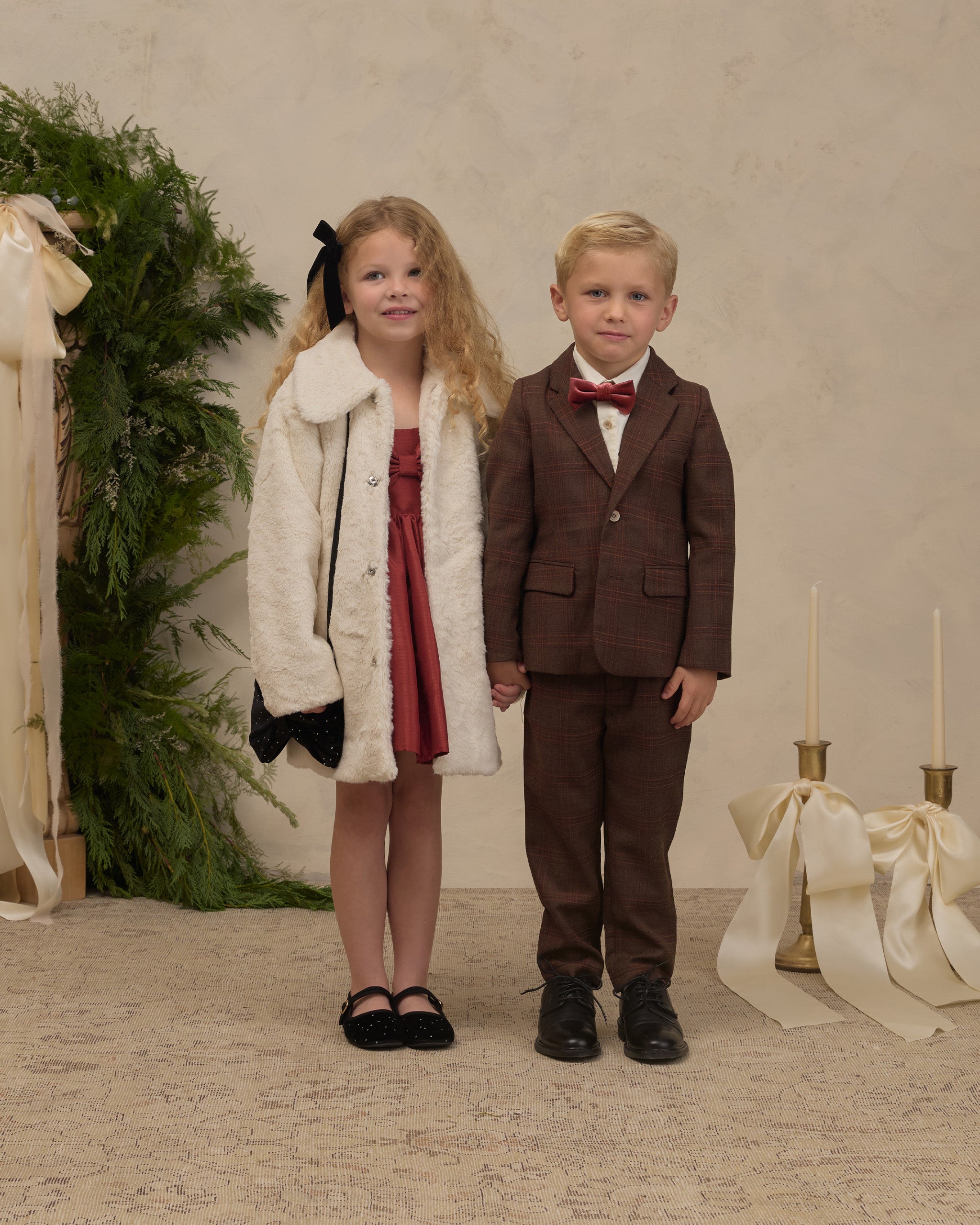 Young girl in a natural faux fur coat stands beside a boy in a brown suit at a festive holiday display for Noralee children's outerwear