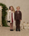 Young girl in a natural faux fur coat stands beside a boy in a brown suit at a festive holiday display for Noralee children's outerwear