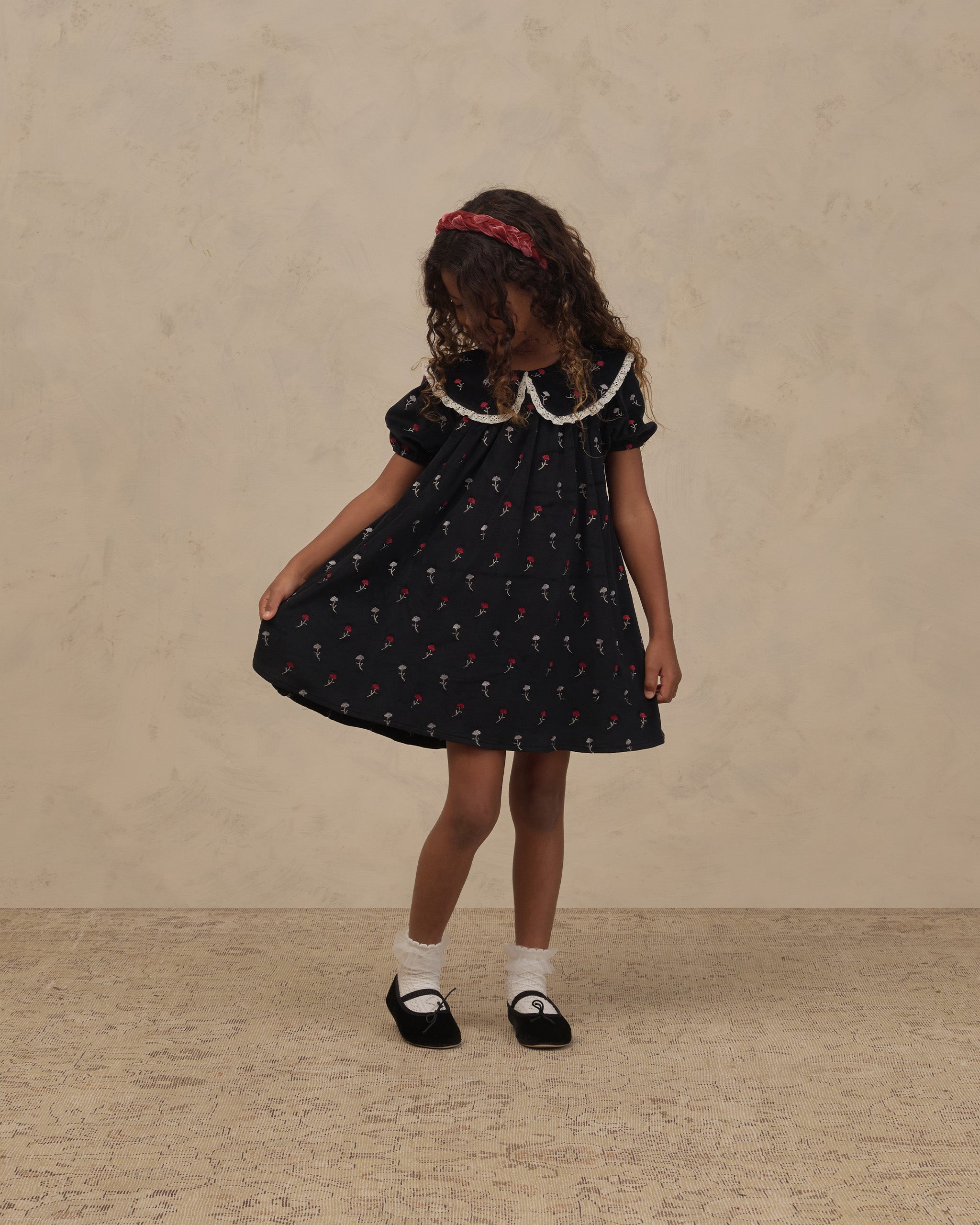 Young girl wearing a Noralee black floral dress with a lace trimmed round collar puff sleeves and full skirt with white socks and Mary Jane shoes
