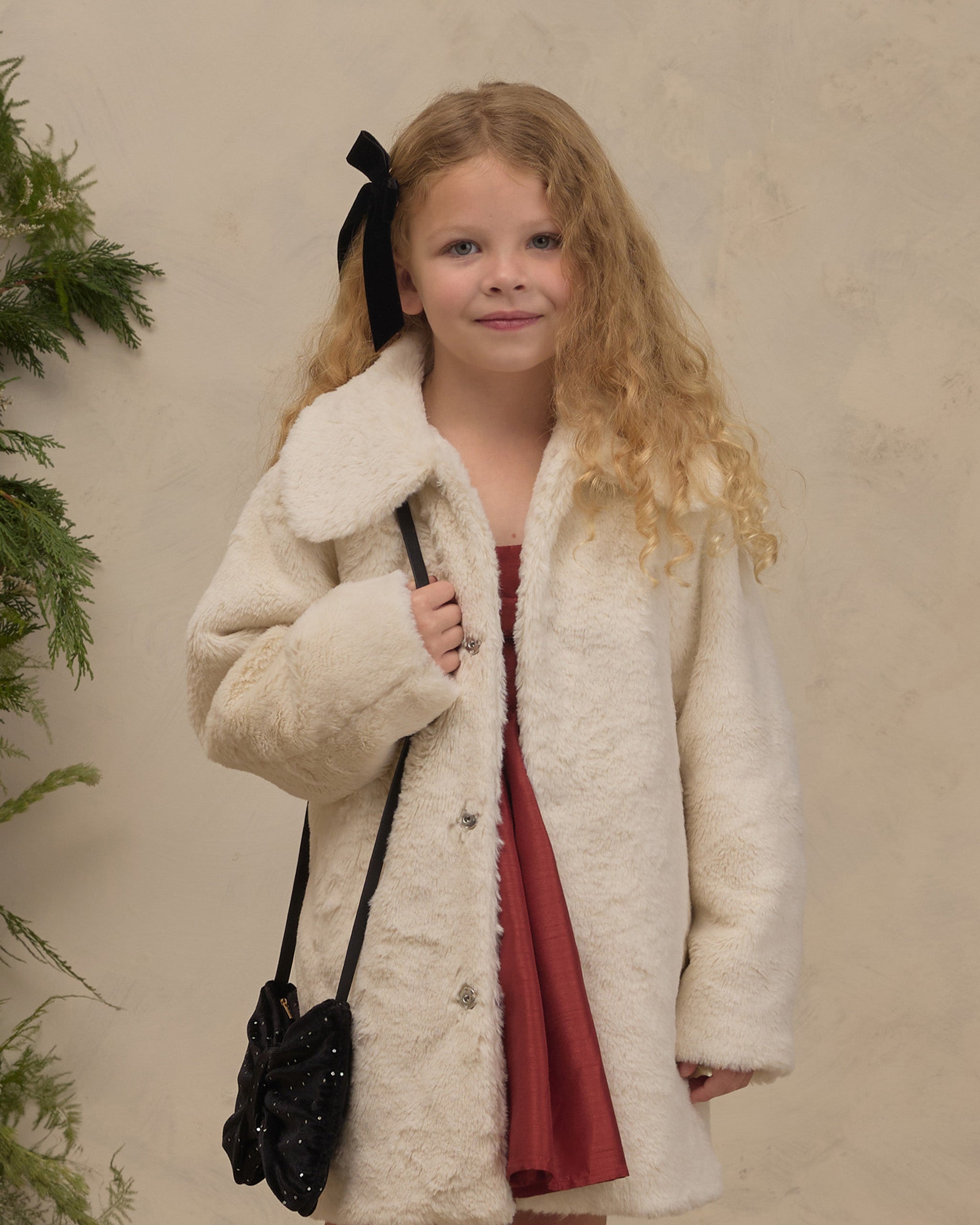 Young girl wearing a cream faux fur coat over a red dress with a black bow and shoulder bag in a cozy holiday setting by Noralee