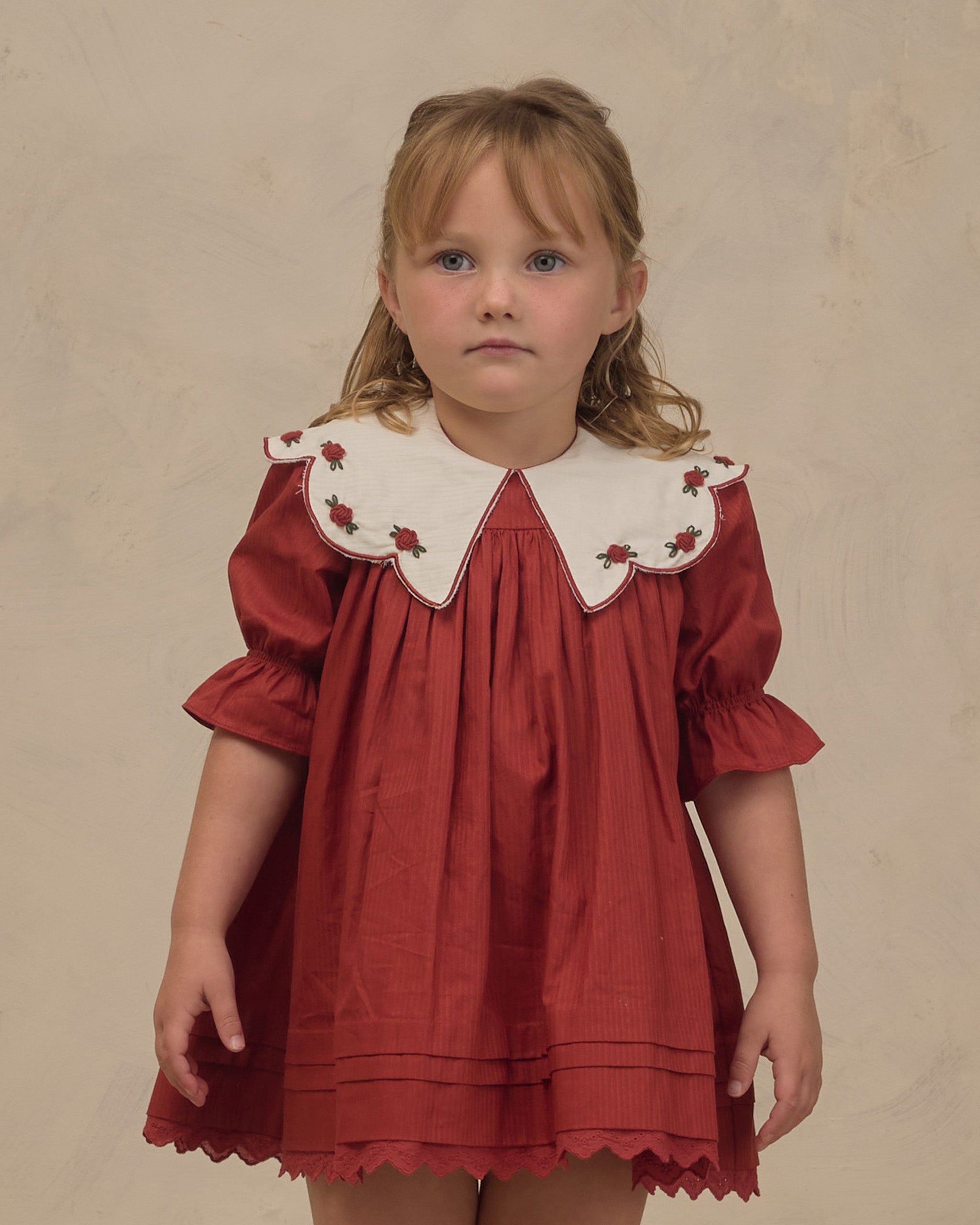 Young girl wearing a ruby red Noralee dress with a scalloped white embroidered collar puff sleeves pleated details and a delicate scalloped hem