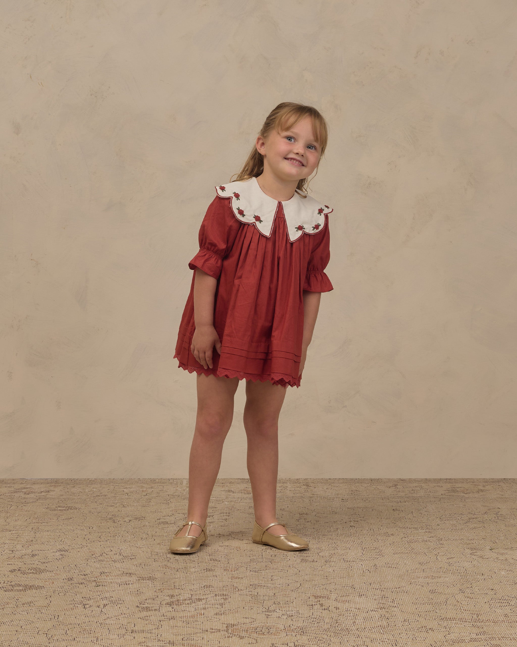 Young girl wearing a ruby red Noralee dress with a white scalloped collar and pleated skirt while standing in gold shoes against a neutral backdrop
