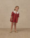 Young girl wearing a ruby red Noralee dress with a white scalloped collar and pleated skirt while standing in gold shoes against a neutral backdrop