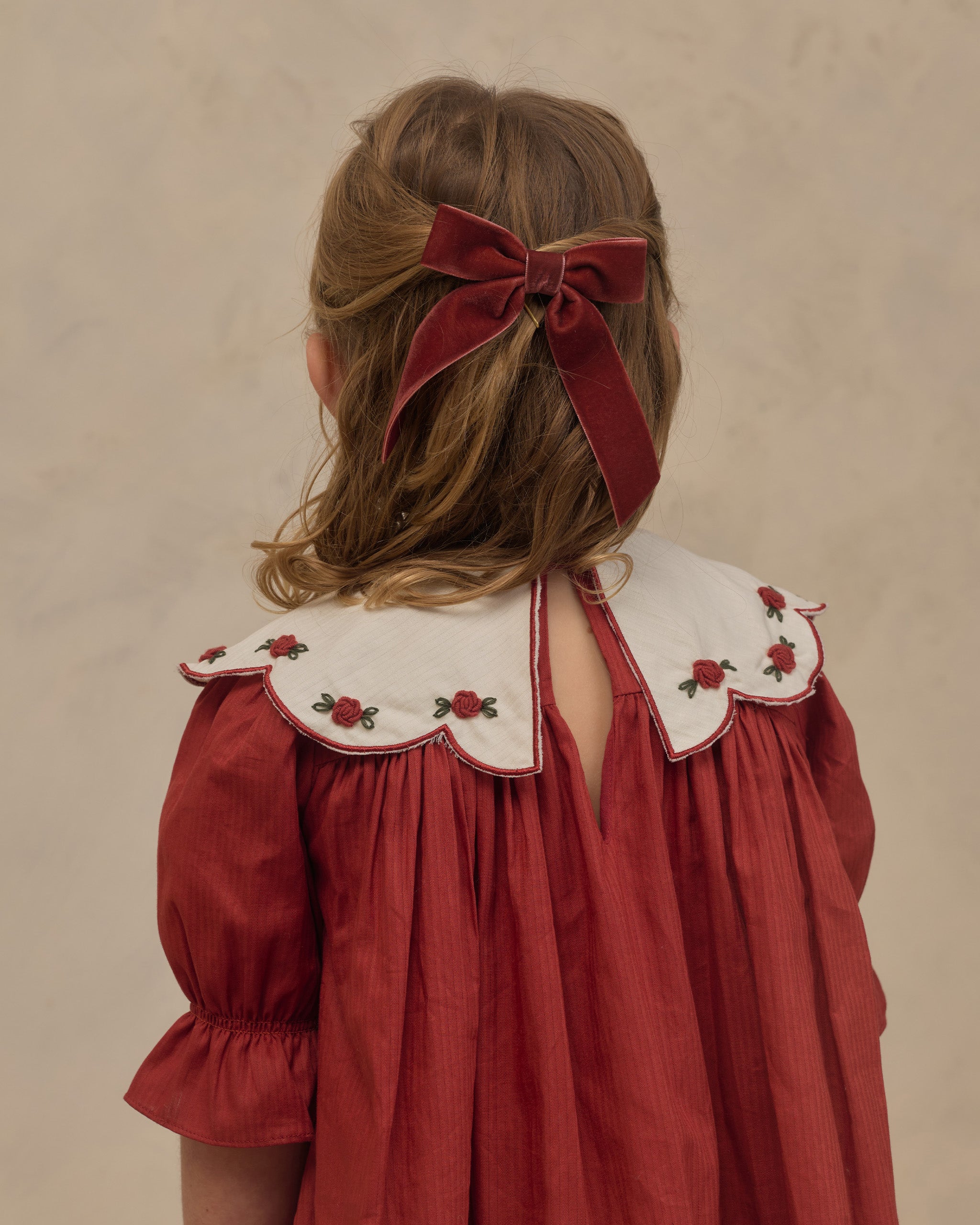 Child seen from behind wearing a ruby red dress with embroidered scalloped collar and puff sleeves with a matching velvet bow in her hair by Noralee