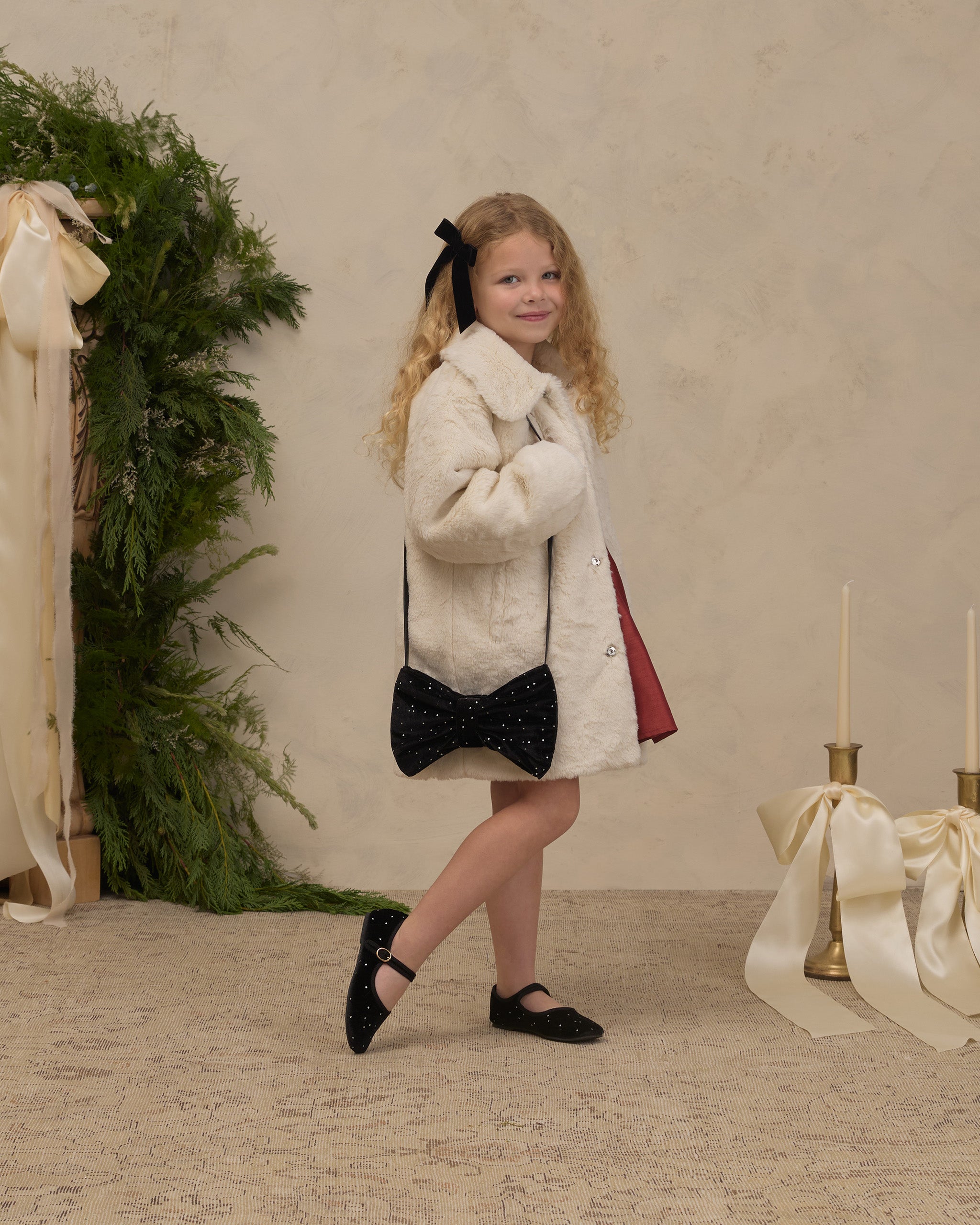 Noralee cream faux fur coat on a young girl with a full collar and hidden front snaps styled with a black bow purse and Mary Jane shoes in a festive room