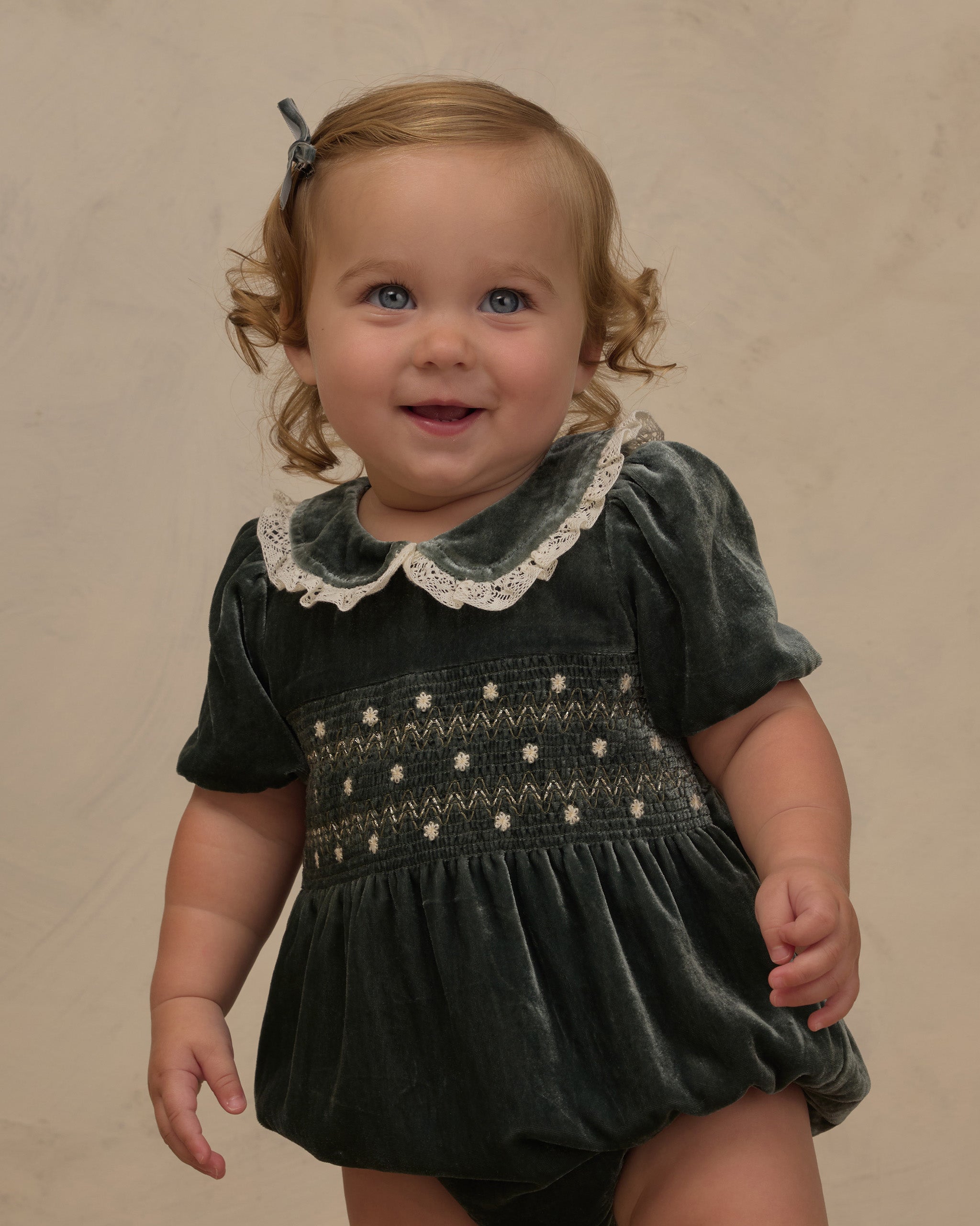 A little girl wearing a green velvet romper with delicate embroidered details.