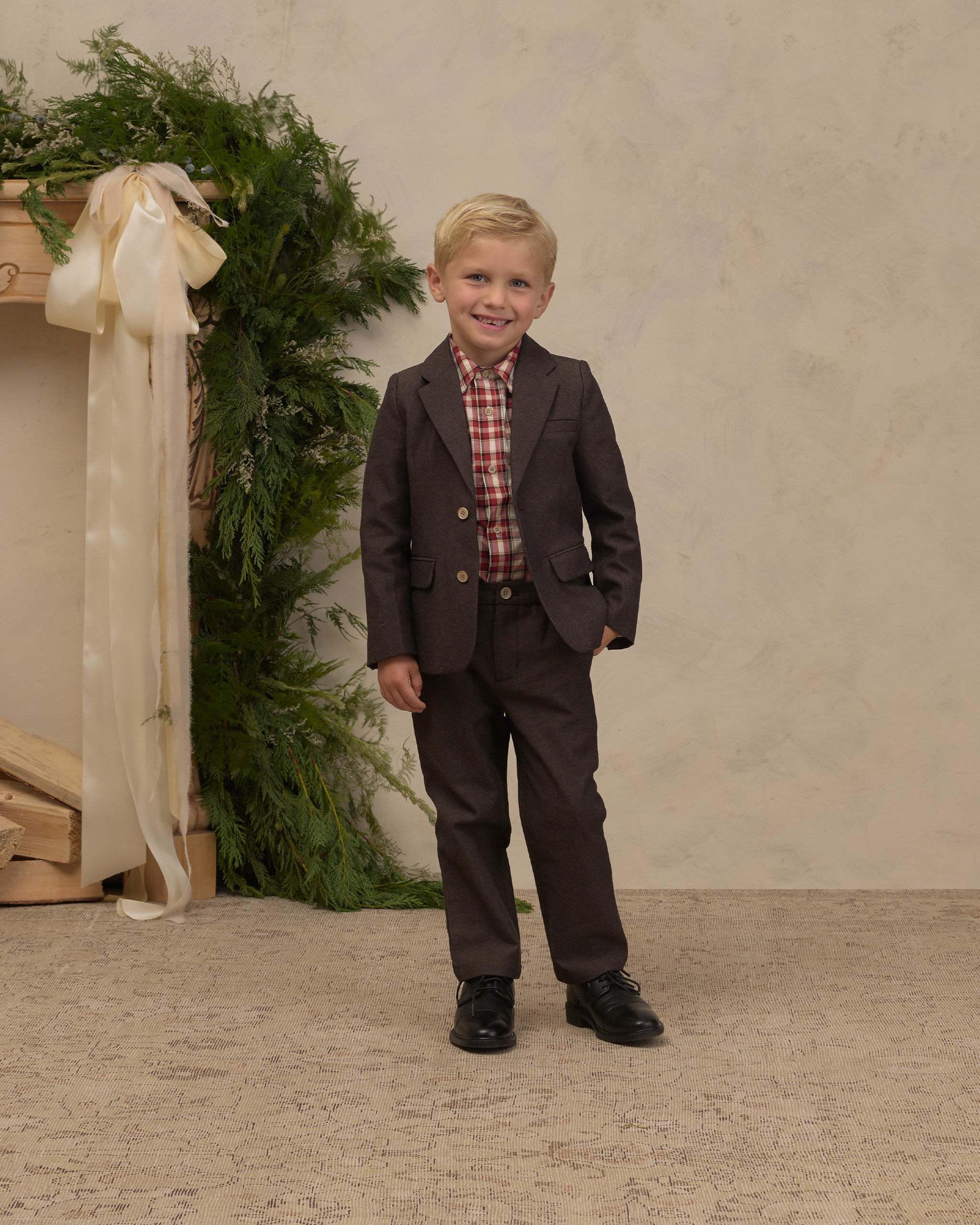 Young boy wearing a Noralee dark brown blazer with notched lapels and front pockets over a red plaid shirt with matching trousers and black shoes