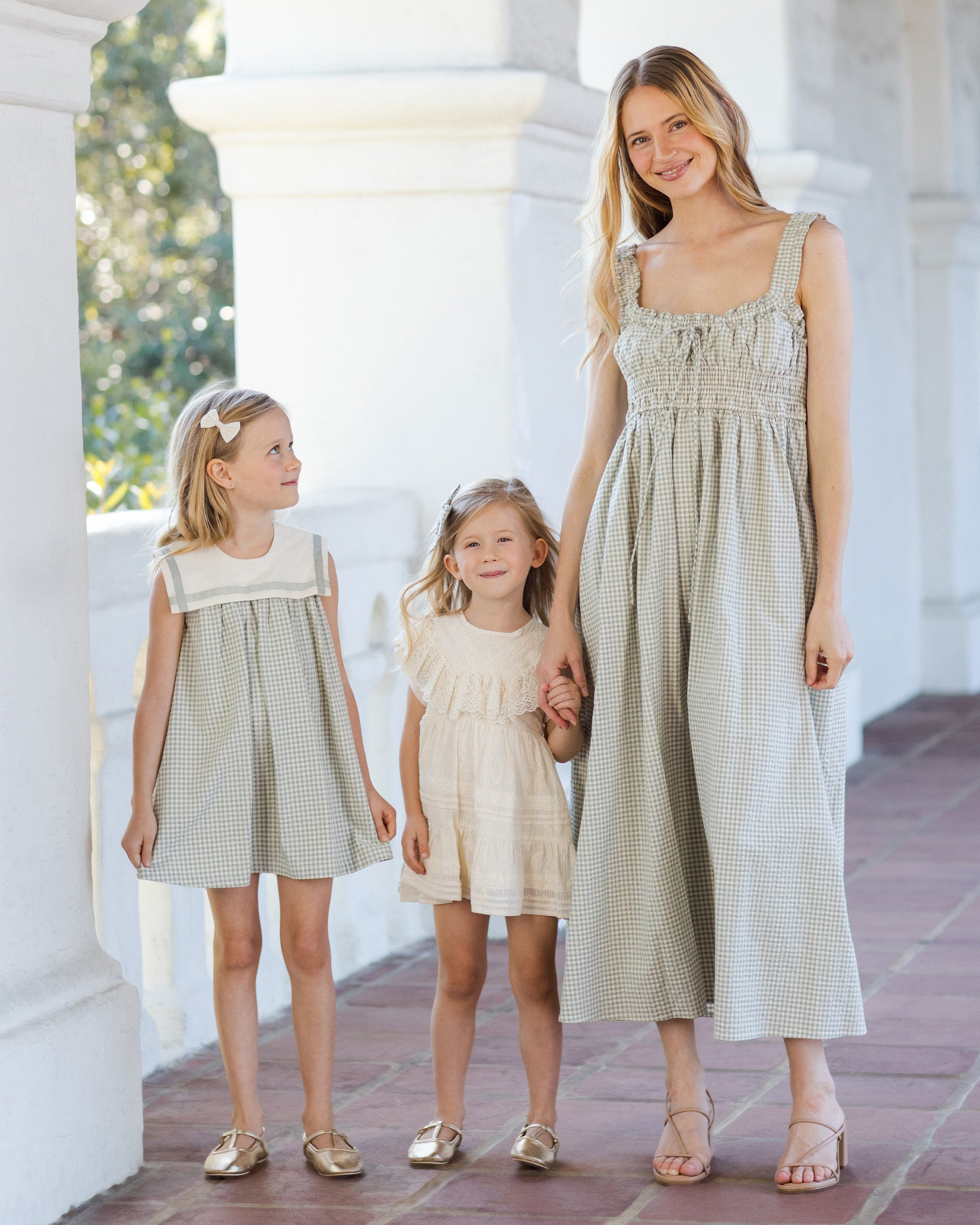 Woman and two young girls in matching dresses standing on a sunlit outdoor path.