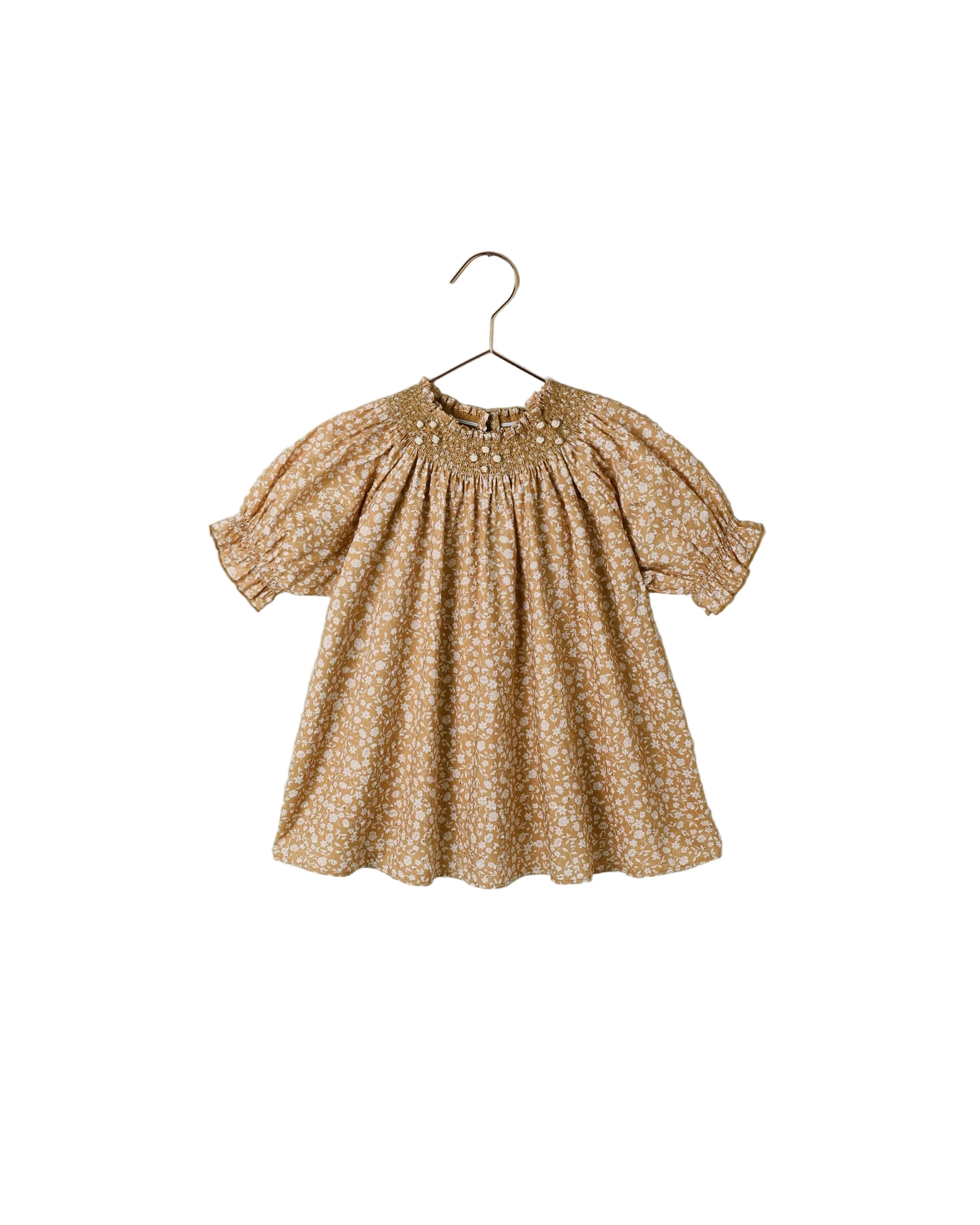 A toddler girl's short-sleeve dress with a smocked bodice and a small white floral print on a warm tan background, hanging on a metal hanger against a white background.