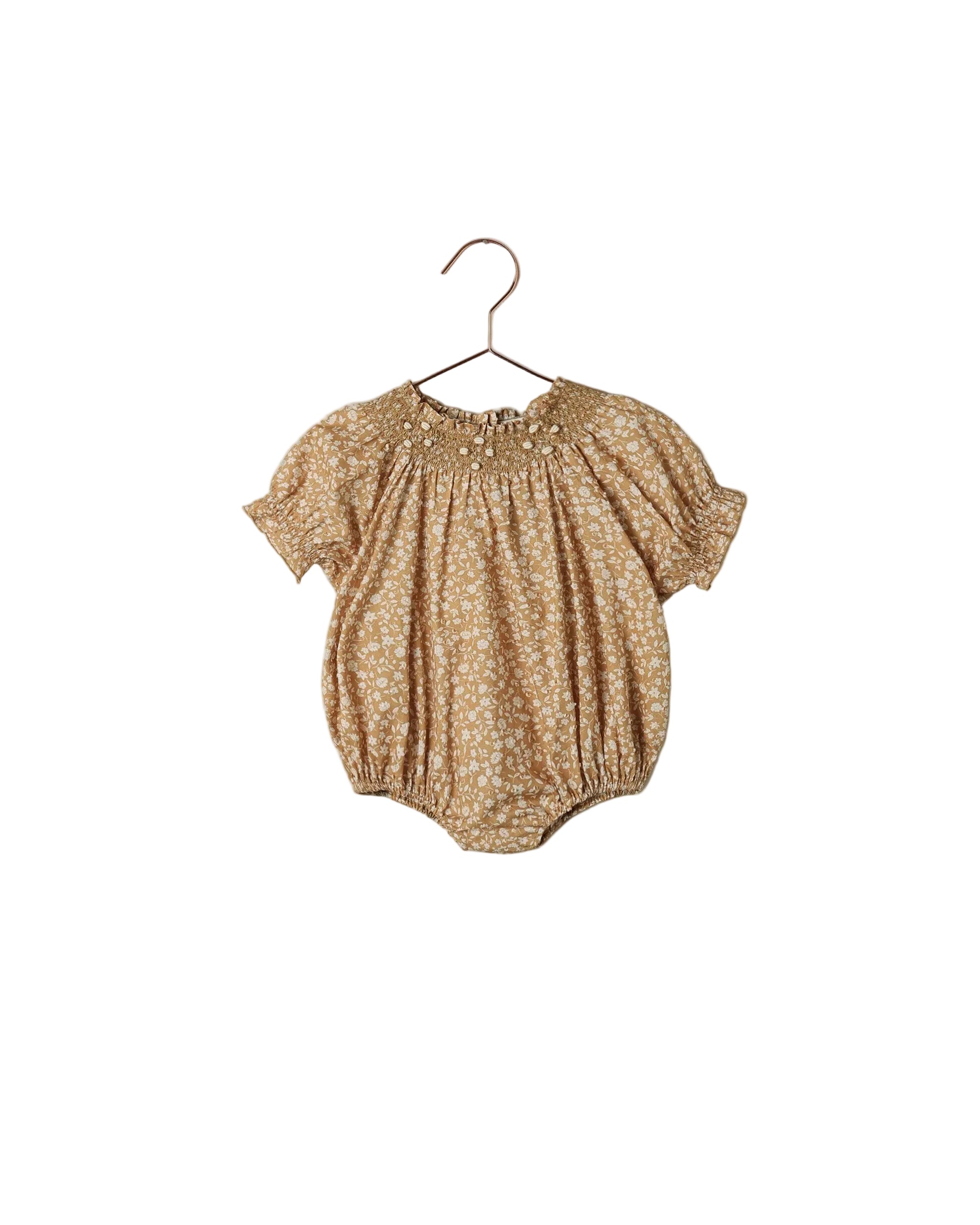 A baby short-sleeve romper in a warm tan and white floral print, hanging on a metal hanger against a white wall.