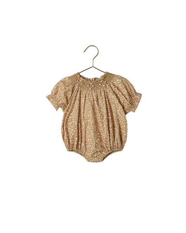 A baby short-sleeve romper in a warm tan and white floral print, hanging on a metal hanger against a white wall.
