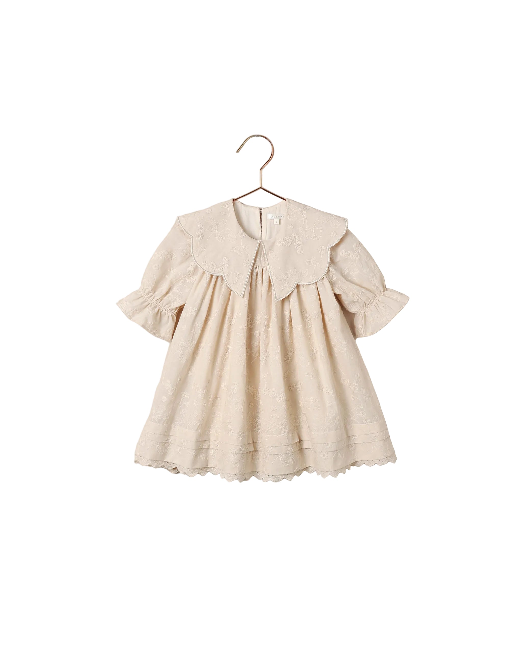 A cream-colored baby dress with a large ruffled Peter Pan collar, short flutter sleeves, and delicate eyelet embroidery, hanging on a wooden hanger against a white backgroundl.