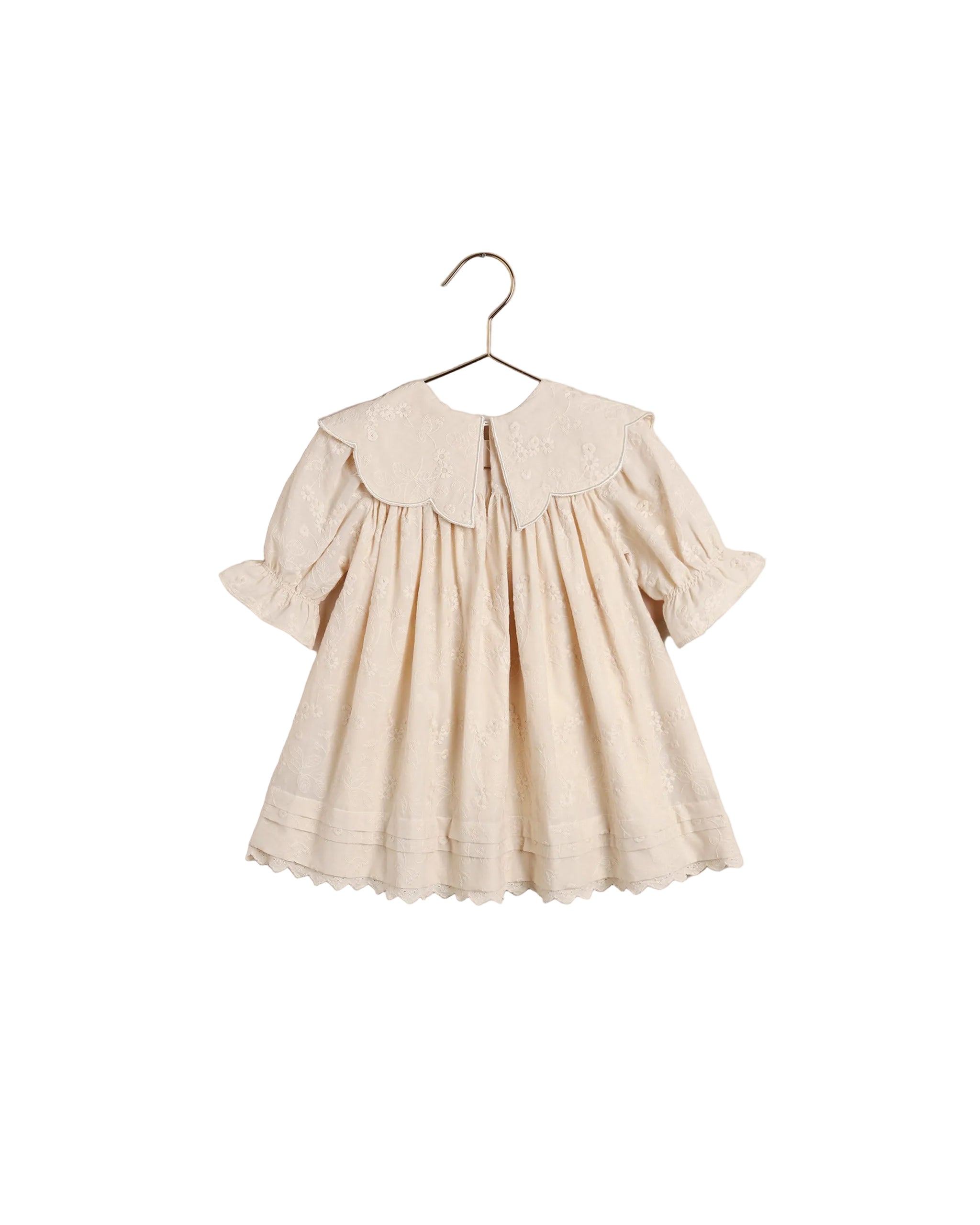 A cream-colored baby dress with a large embroidered Peter Pan collar, short puff sleeves, and a scalloped hem, hanging on a metal hanger against a white backgroundl.