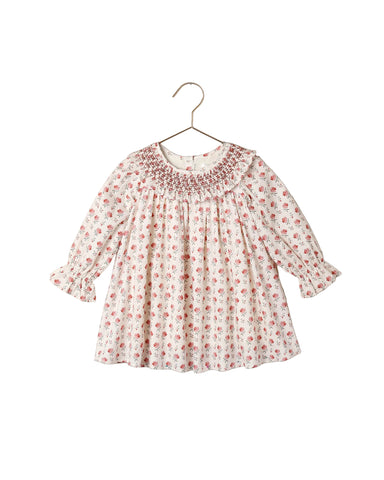 An ivory floral dress with a  delicate smocked collar and long sleeves with elastic cuffs to its full, gathered body. The back has a shell button placket at the back for easy changes.