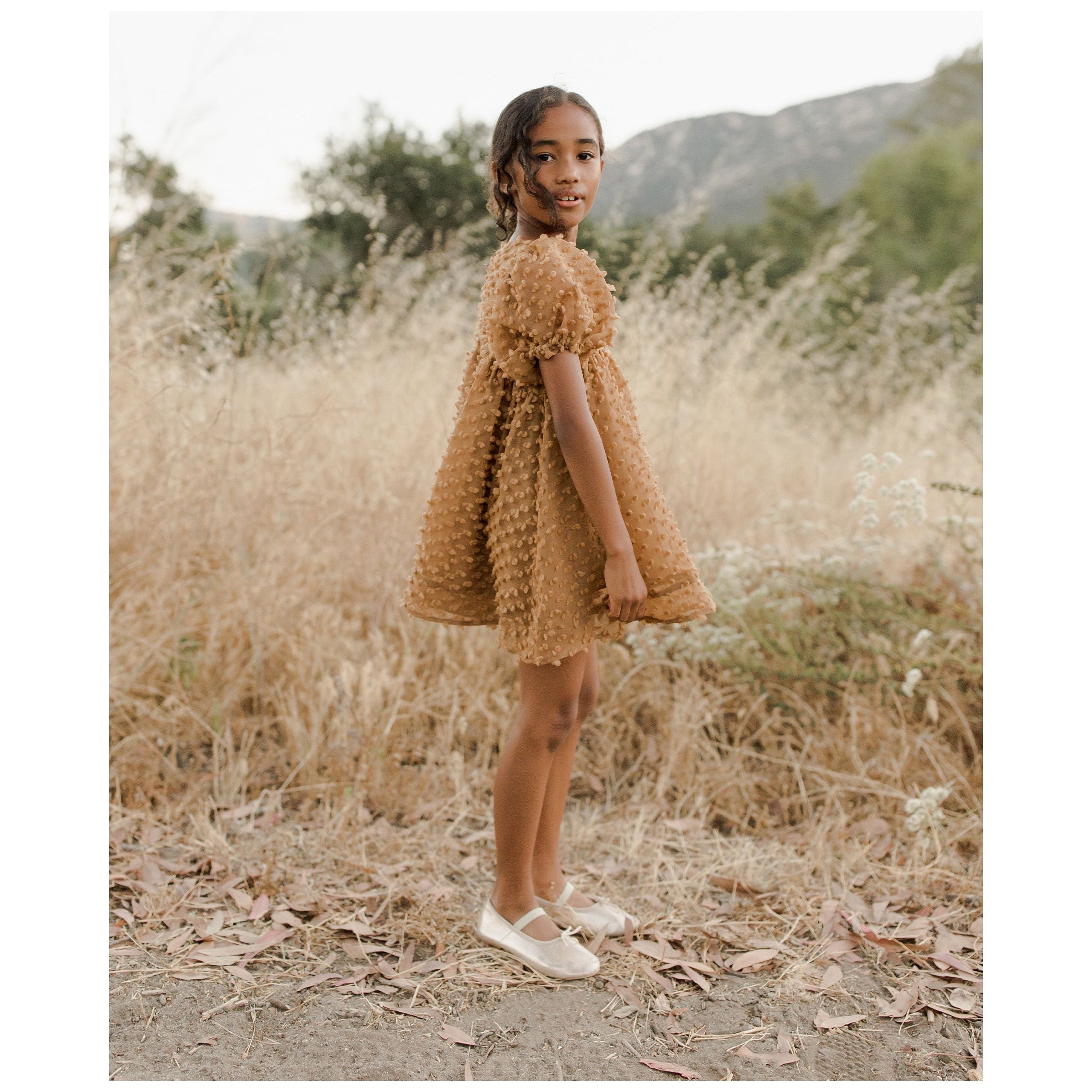 girl wearing gold colored babydoll style dress with popcorn fabric and puff sleeves with gold ballet flats