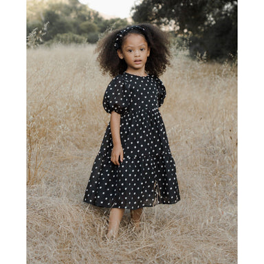 girl wearing black midi dress with layered tiered skirt, full puffed sleeves, and a delicate button-back closure and white polka dots