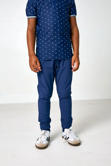 A child wearing a navy blue short-sleeve polo shirt with a white polka dot pattern, navy blue joggers, and white sneakers, standing against a plain white studio background.