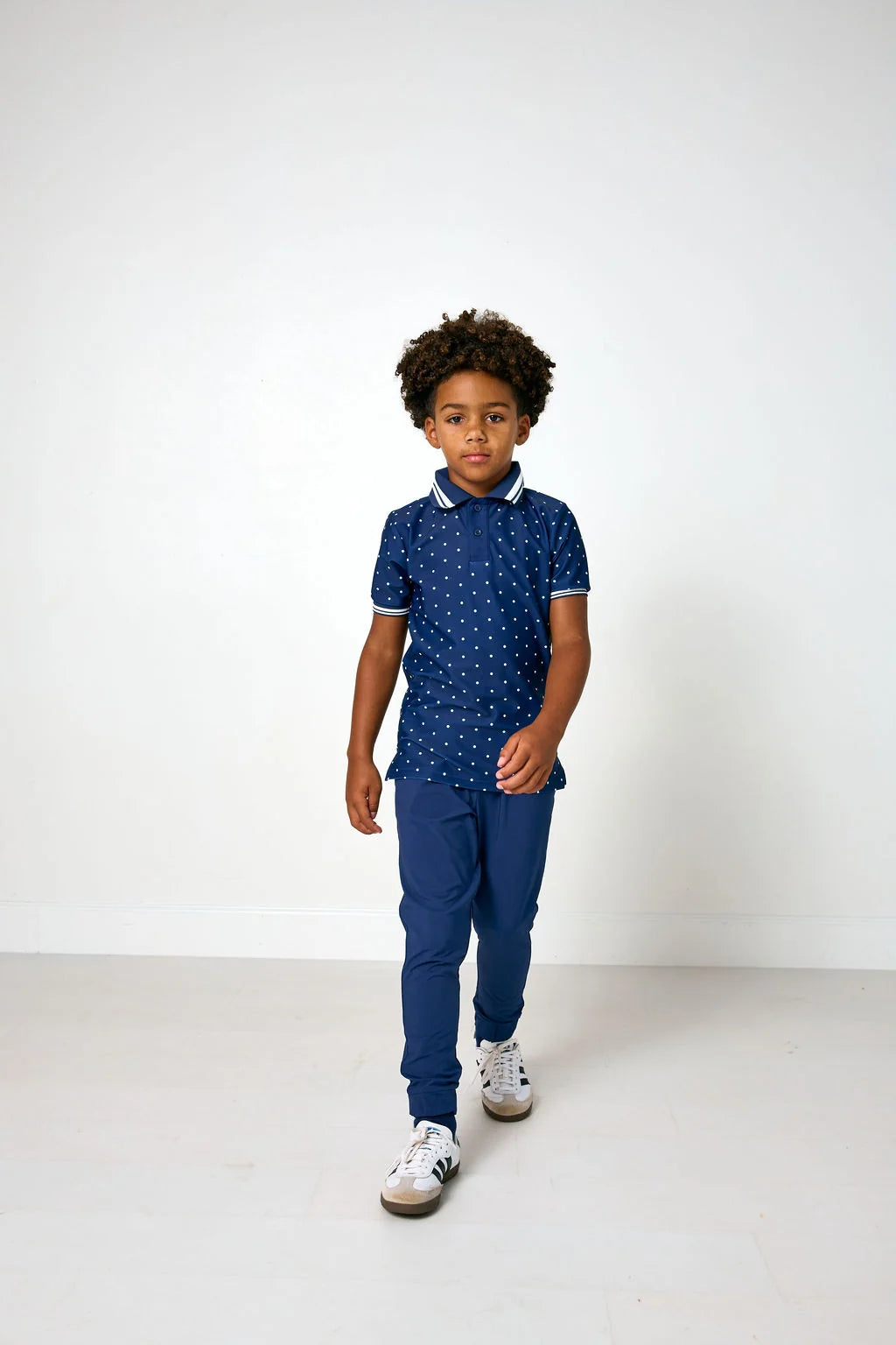 A young boy walking forward in a studio setting, wearing a navy blue short-sleeve polo shirt with a small white pattern, navy joggers, and white sneakers.