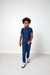 A young boy walking forward in a studio setting, wearing a navy blue short-sleeve polo shirt with a small white pattern, navy joggers, and white sneakers.