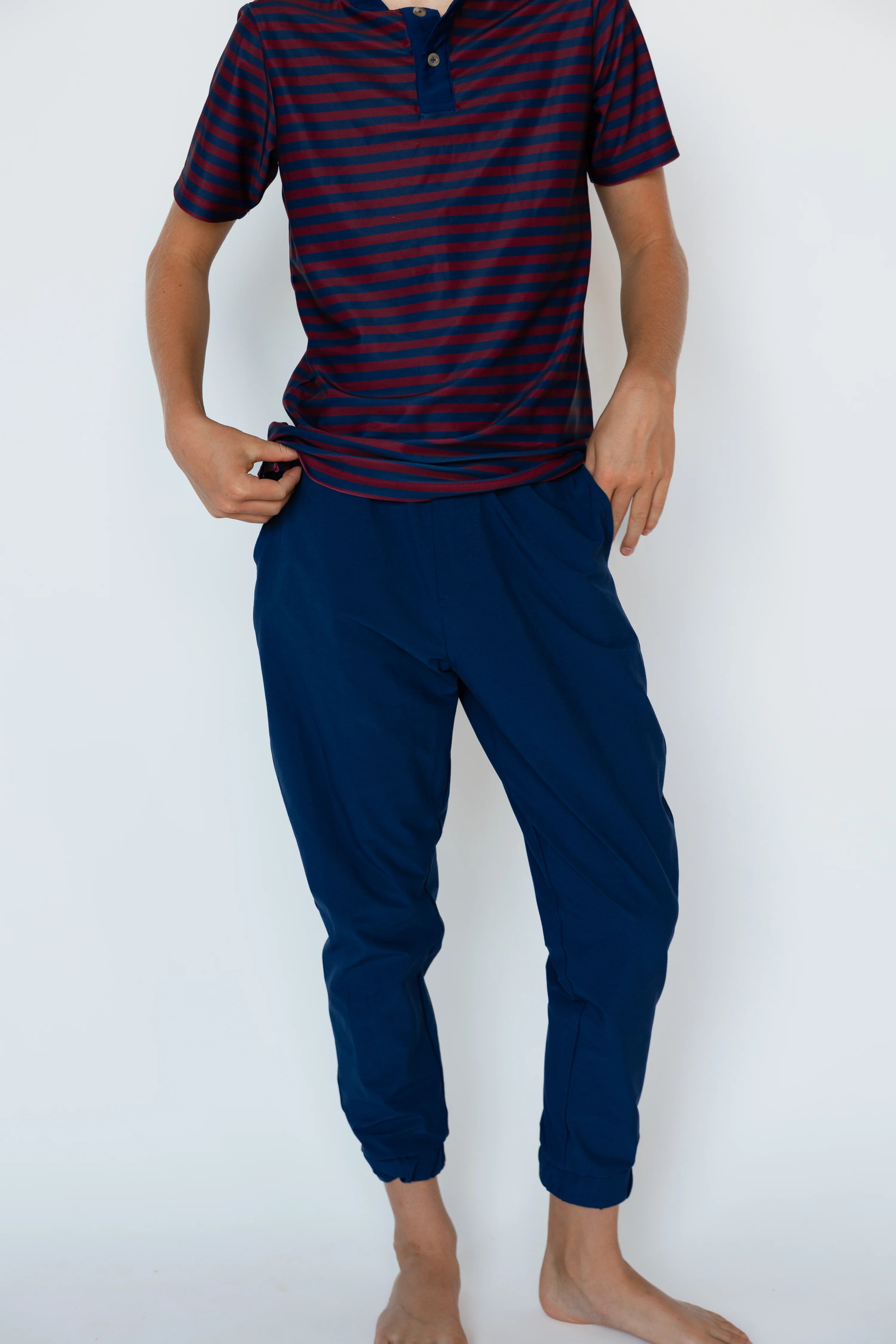 A man wearing a navy blue and red striped short-sleeve polo shirt with dark navy jogger pants, standing barefoot against a plain white background.