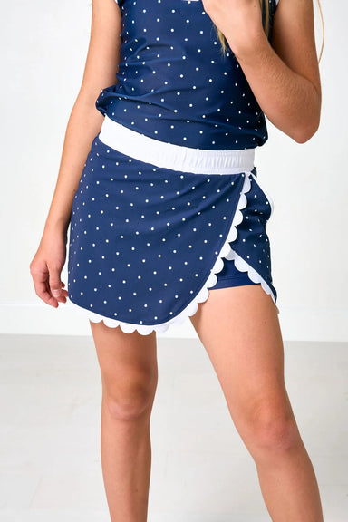 A model wearing a navy blue and white polka dot mini skort with a white waistband and scalloped edges, paired with a matching top.