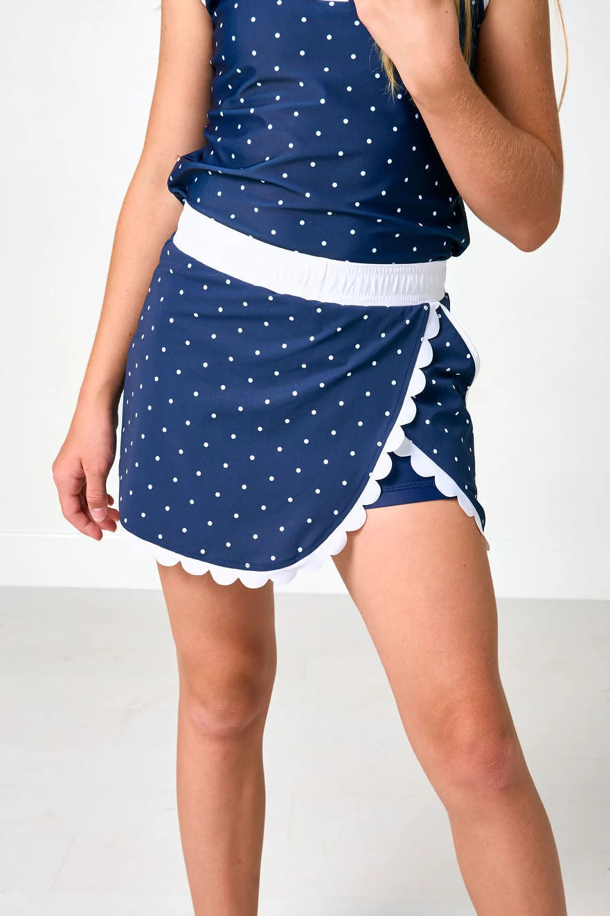 A model wearing a navy blue and white polka dot mini skort with a white waistband and scalloped edges, paired with a matching top.