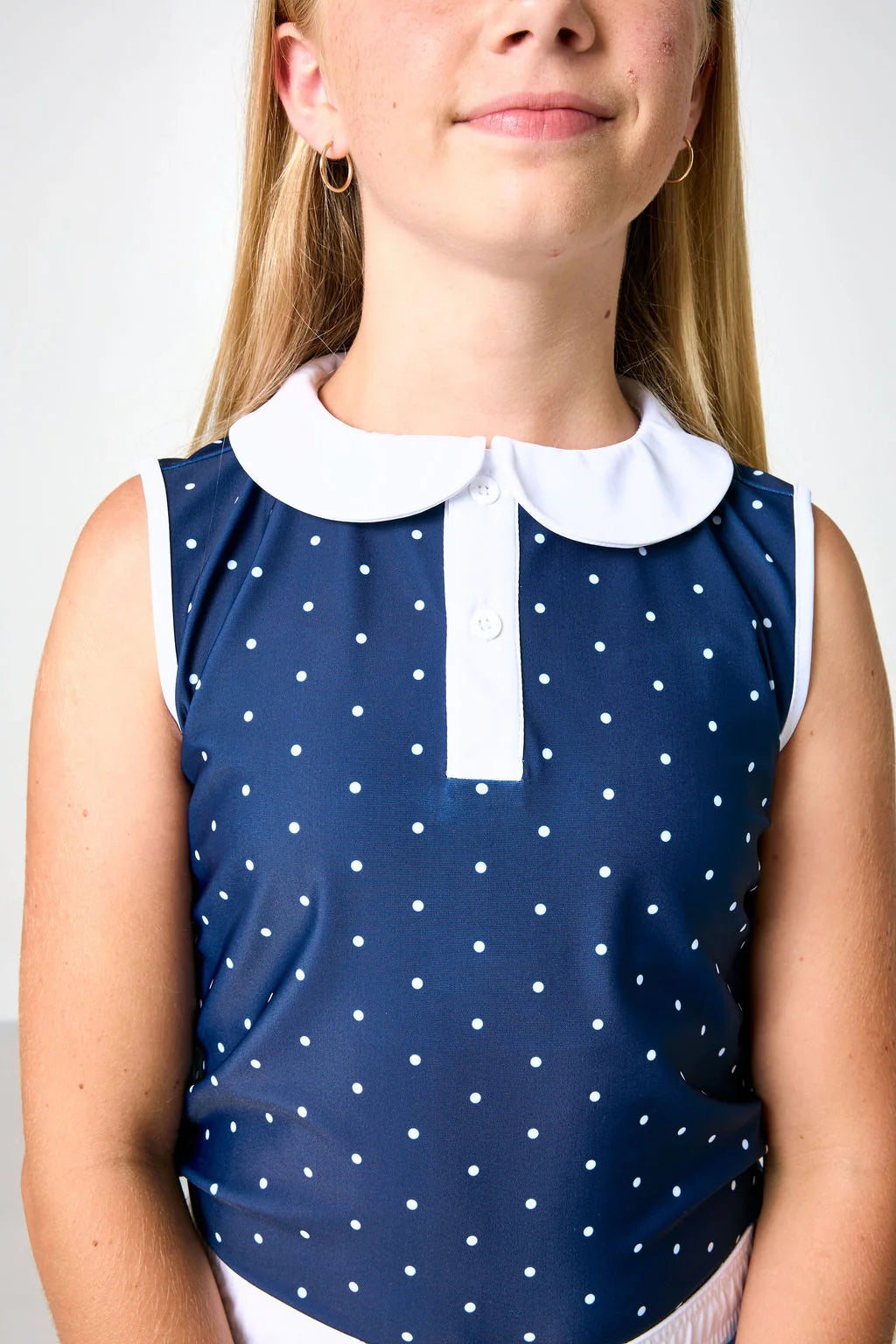 A close-up of a child wearing a navy sleeveless top with white polka dots and a white Peter Pan collar.
