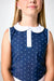 A close-up of a child wearing a navy sleeveless top with white polka dots and a white Peter Pan collar.