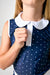 Close-up of a woman wearing a navy blue sleeveless top with white polka dots and a contrasting white Peter Pan collar.