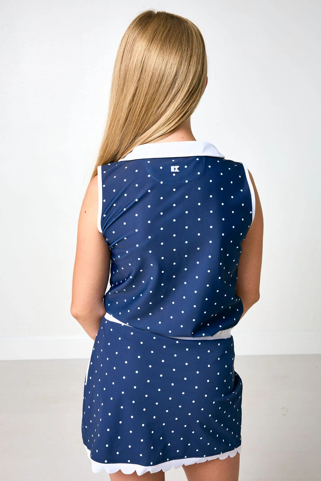 A young girl viewed from behind wearing a sleeveless navy blue dress with a white polka dot pattern and white trim, set against a plain white studio background.