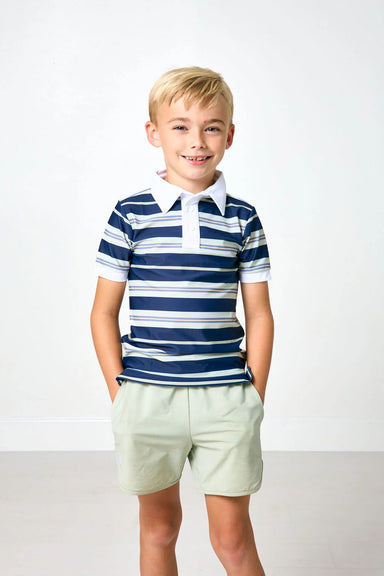 A young boy with blonde hair stands against a white studio background wearing a navy blue and white striped short-sleeve polo shirt with light green shorts.