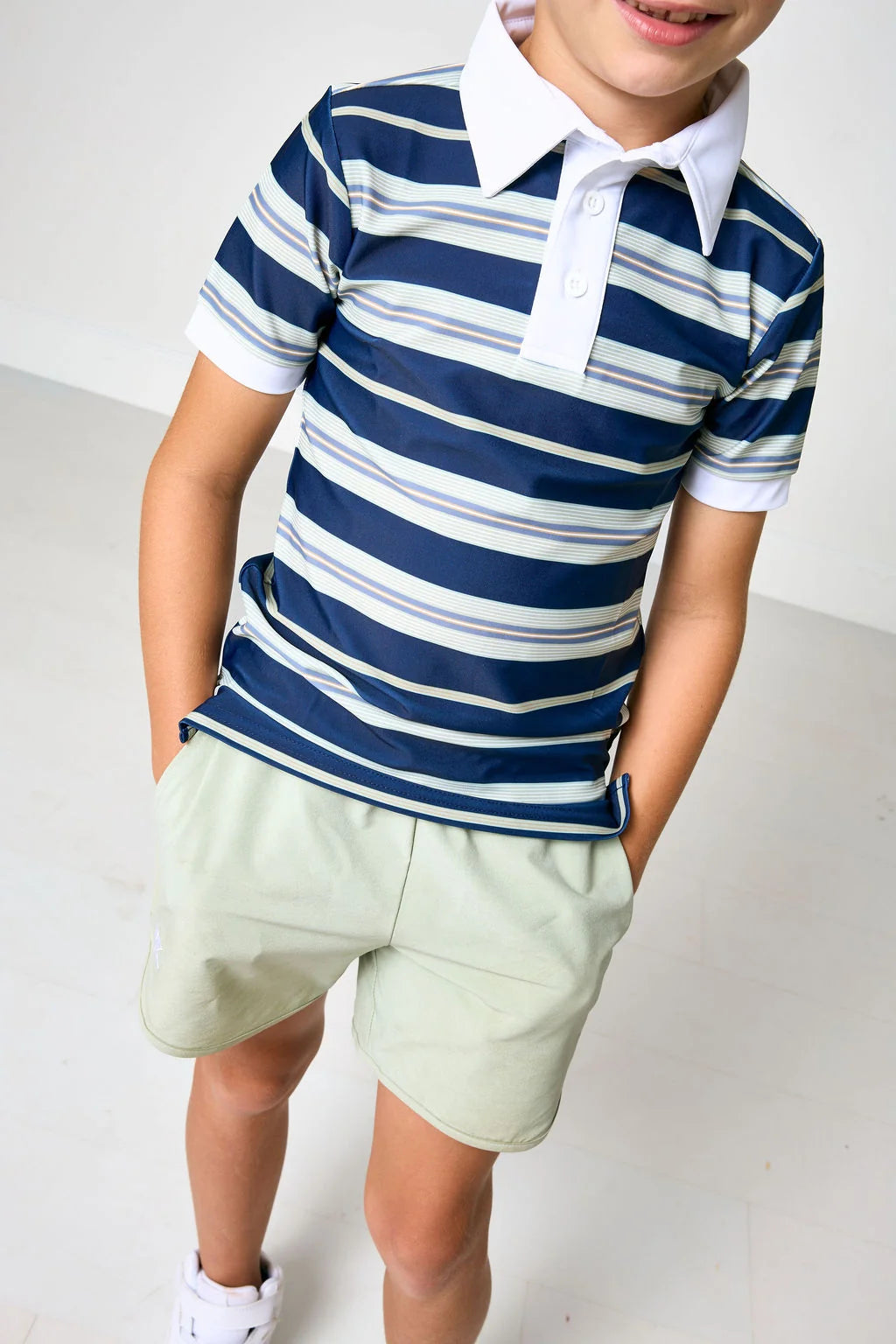 A child wearing a navy blue and white striped short-sleeve polo shirt with a white collar, paired with light green shorts, standing against a plain light gray background.