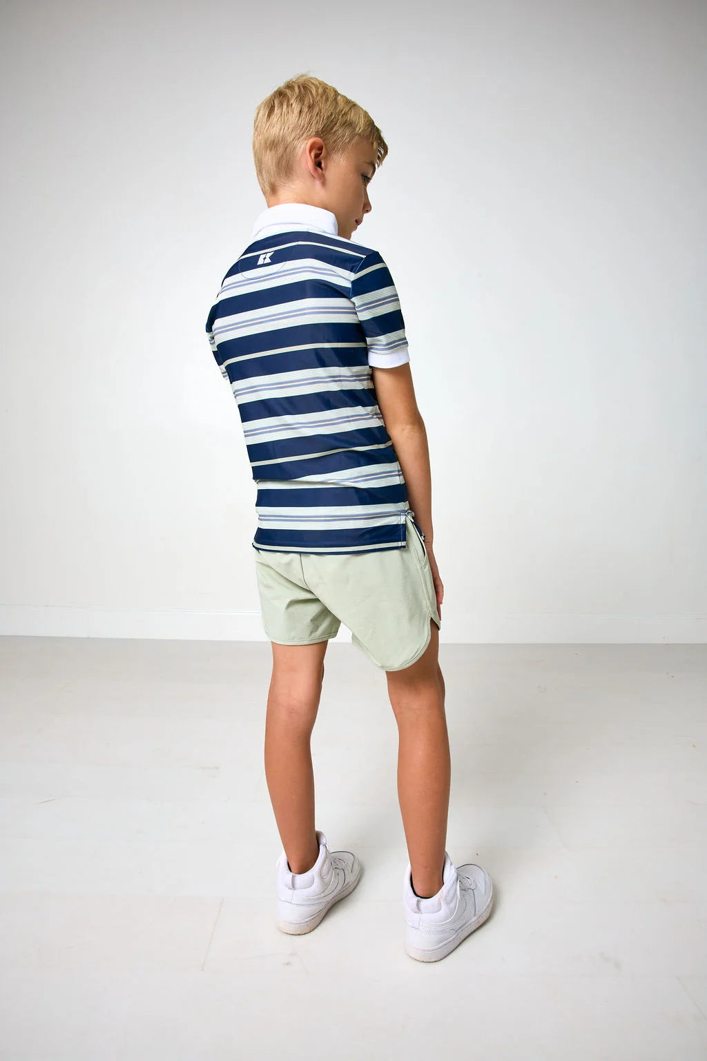 A young boy with blonde hair is shown from behind wearing a navy blue and white striped short-sleeve polo shirt, light green shorts, and white sneakers, standing against a plain white studio background.