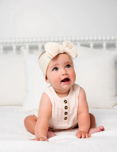 A baby sitting on a white bed wearing a cream-colored sleeveless romper with gold buttons and a matching cream bow headband.