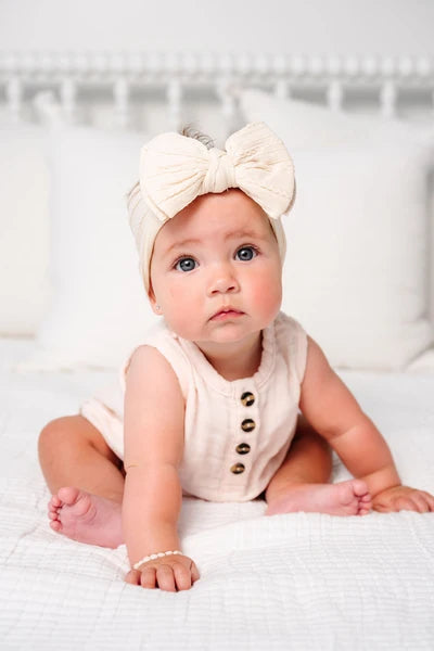 A baby sitting on a white bed wearing a cream-colored sleeveless romper with gold buttons and a matching cream bow headband.