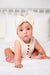 A baby sitting on a white bed wearing a cream-colored sleeveless romper with gold buttons and a matching cream bow headband.