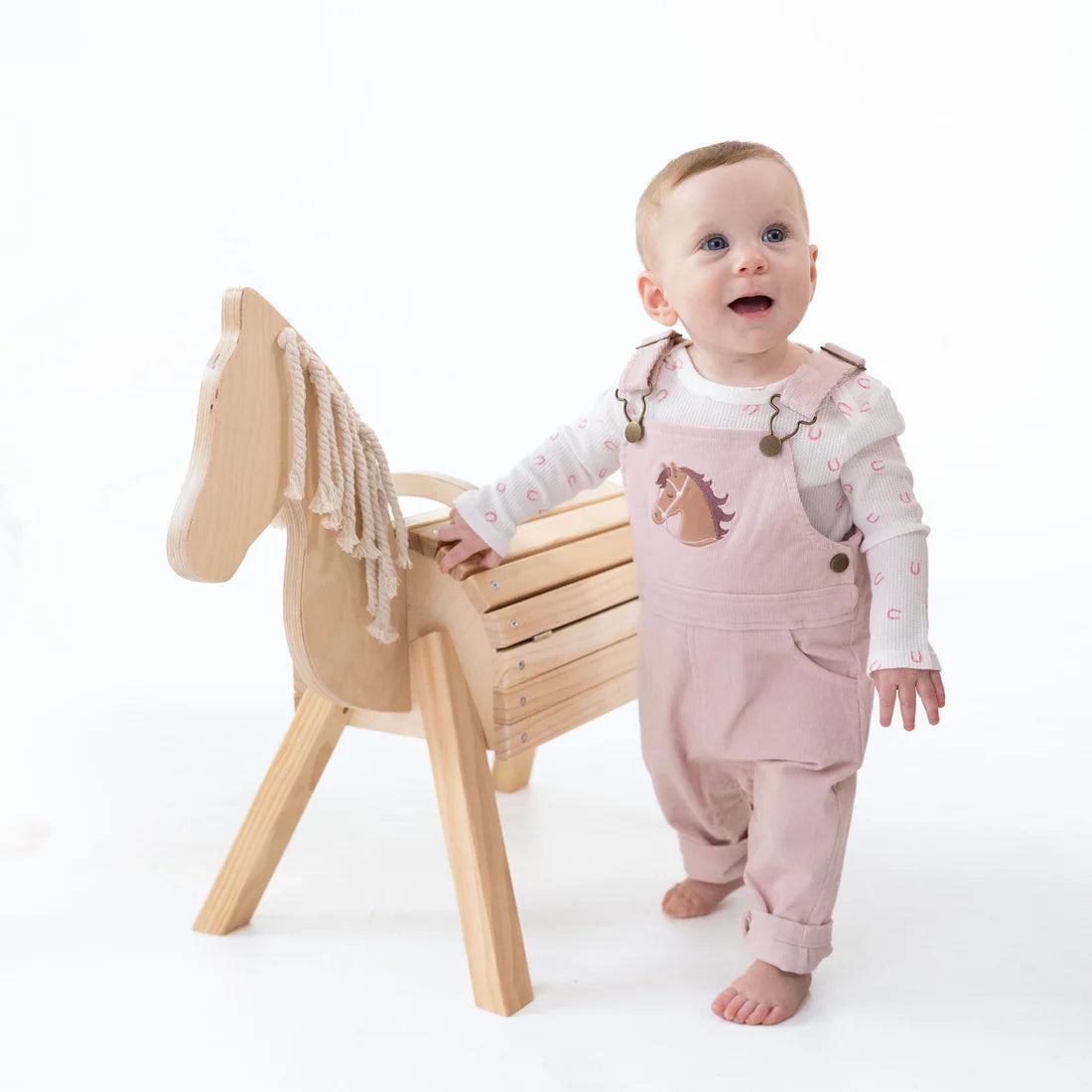 A baby standing barefoot next to a wooden rocking horse, wearing a light pink overall outfit with a horse print on the chest and a white long-sleeve shirt with small pink hearts.
