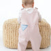 A baby seen from behind wearing a light pink corduroy overall romper over a white long-sleeve shirt, standing on a round beige textured pouf against a white background.