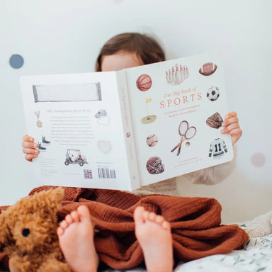 Child reading a book titled 'Big Book of Sports' with a teddy bear on a bed.