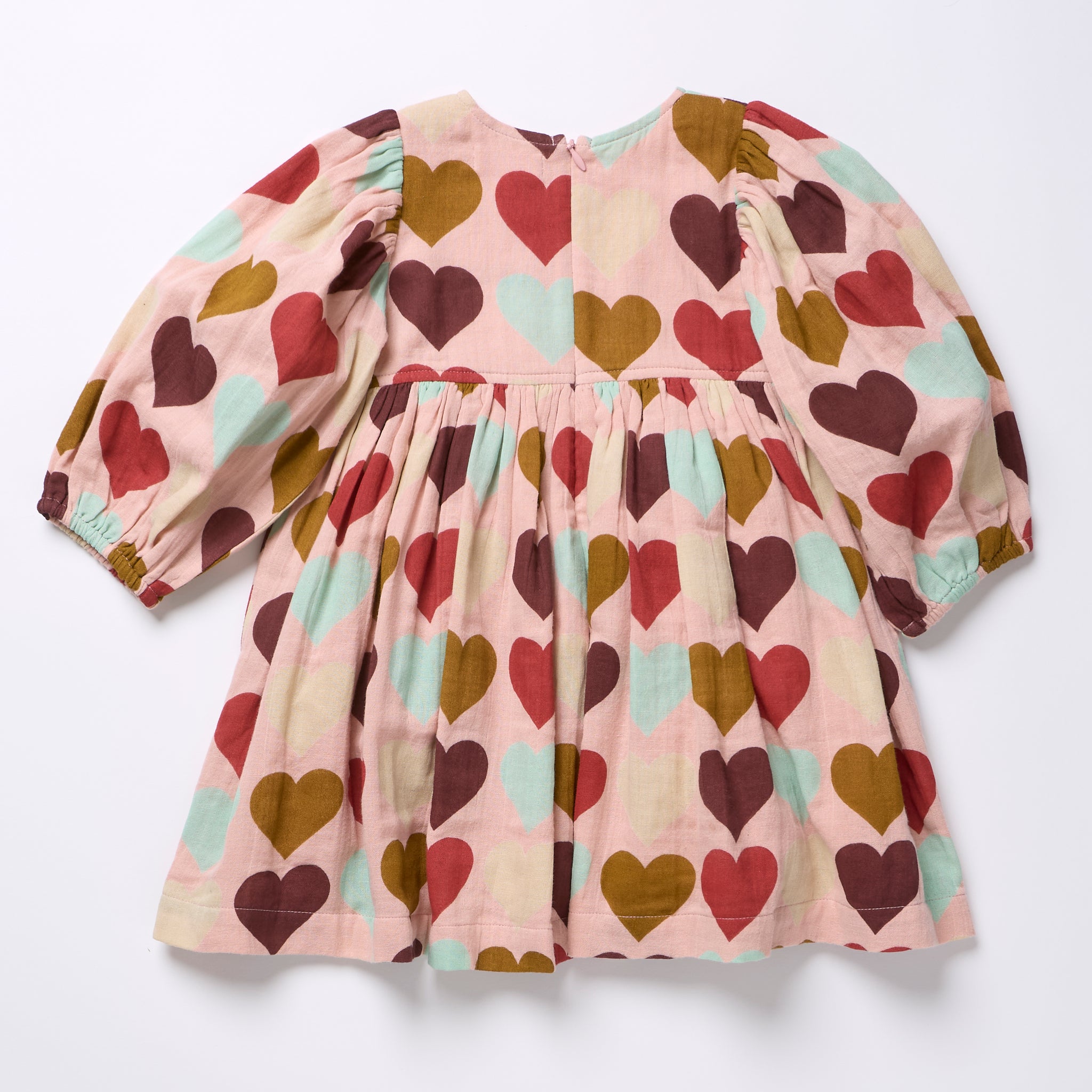 A long-sleeve baby dress with a pink background and an all-over heart pattern in brown, red, and mint green. The dress features puffed sleeves, a gathered waist, and a flared skirt, laid flat on a white background.