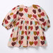 A long-sleeve baby dress with a pink background and an all-over heart pattern in brown, red, and mint green. The dress features puffed sleeves, a gathered waist, and a flared skirt, laid flat on a white background.