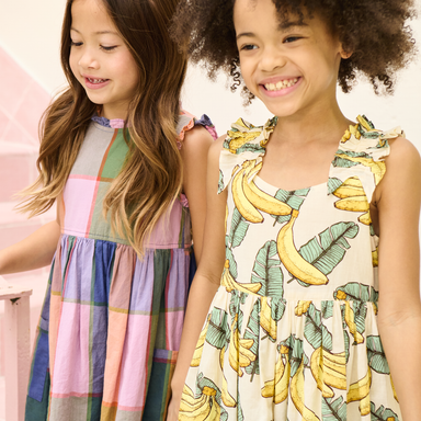 Two young girls wearing colorful dresses, one with a banana pattern, against a light background.