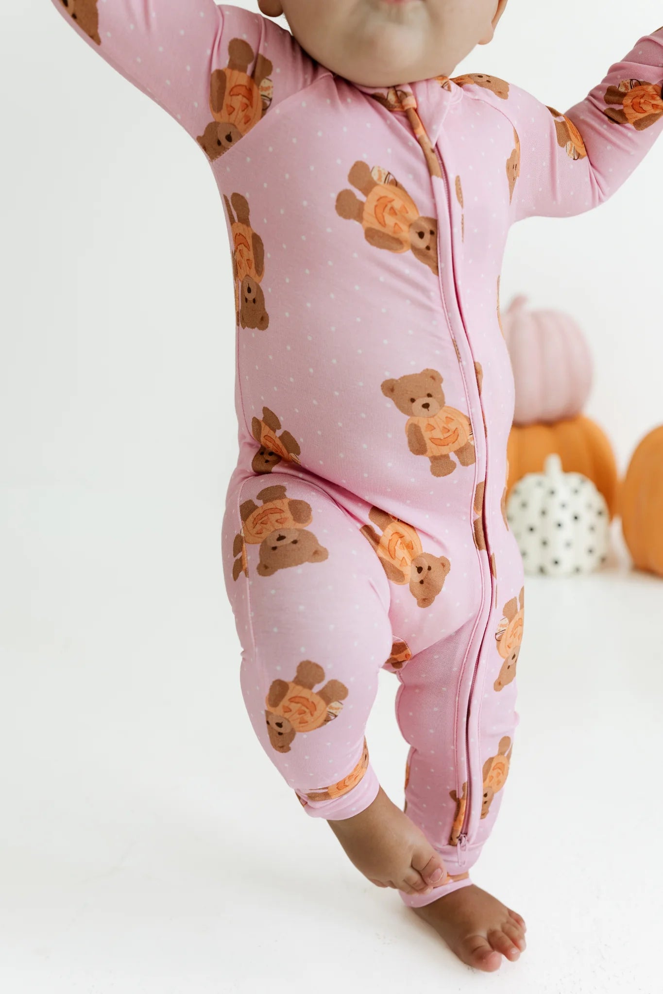 A baby wearing a pink footed zip-up sleeper with an orange and brown teddy bear pattern, shown from the waist down against a white studio background with decorative pumpkins.