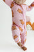 A baby wearing a pink footed zip-up sleeper with an orange and brown teddy bear pattern, shown from the waist down against a white studio background with decorative pumpkins.