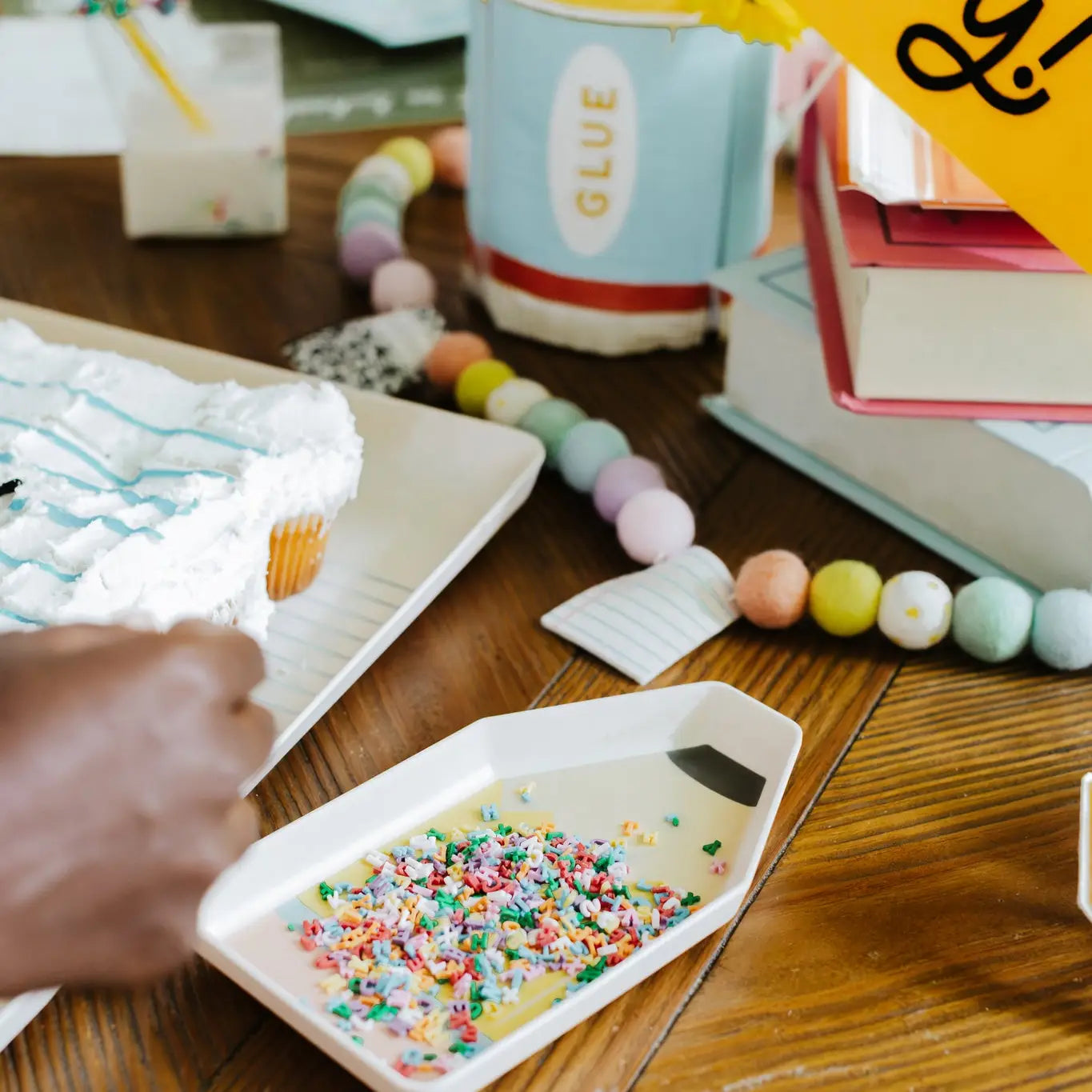 Child's hand reaching for a container of colorful sprinkles on a wooden table with art supplies.