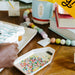 Child's hand reaching for a container of colorful sprinkles on a wooden table with art supplies.